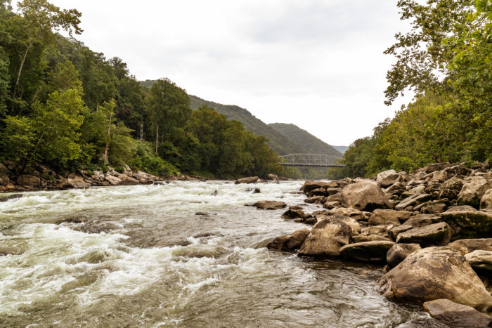 Rushing river with large brown rocks along the shore and a bridge downriver in the distance