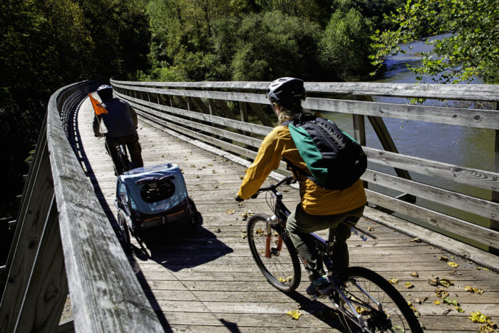 Two bikers on a wooden bridge over a river, one bike towing a small baby carrier